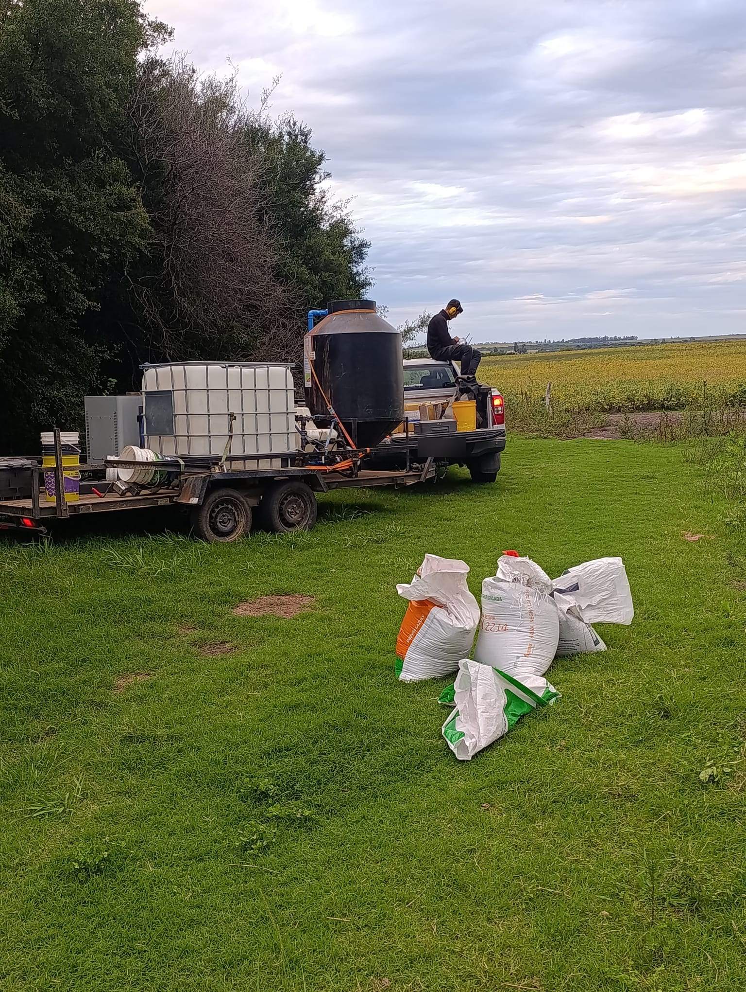 Ein Anhänger mit Wassertank und Mischtechnik steht an einem Feldrand neben Saatgutsäcken.
