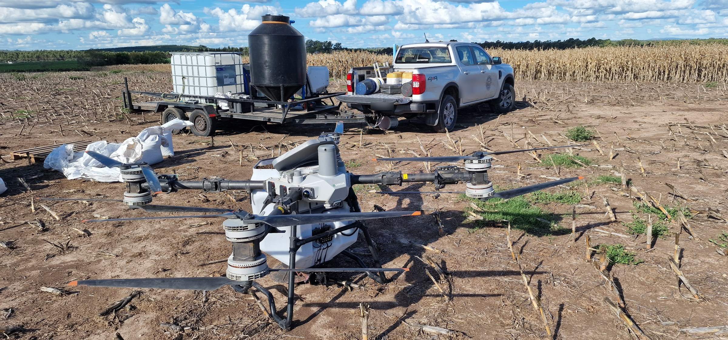 Agrardrohne auf einem abgeernteten Feld, dahinter Anhänger und Pick-up.